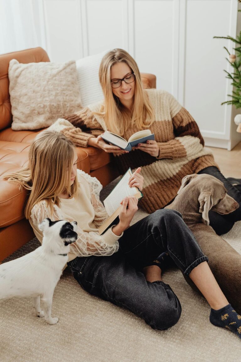 A cozy family scene with mother and daughter reading books together on the sofa with their dogs.